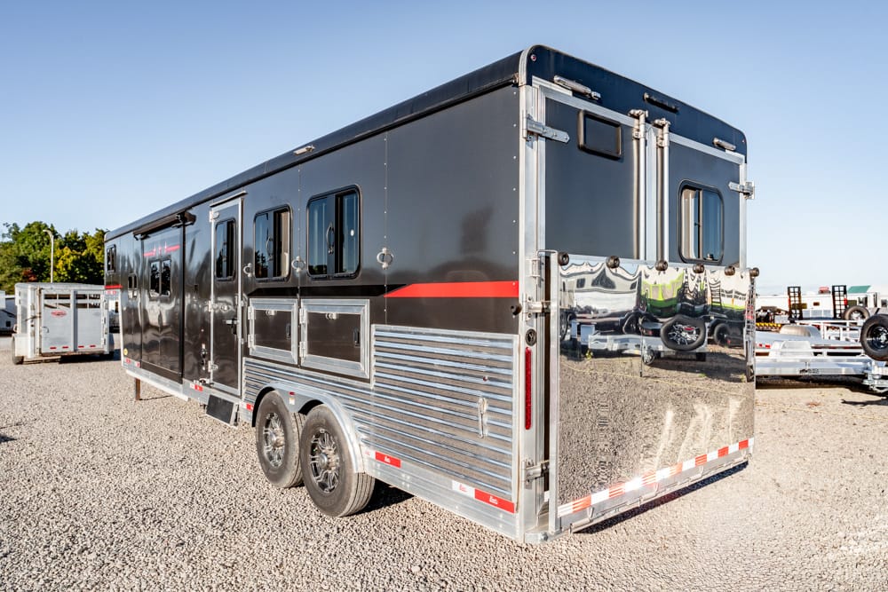 Black and silver horse trailer on a gravel lot, double axle with chrome rear doors and windows, reflecting vehicles nearby