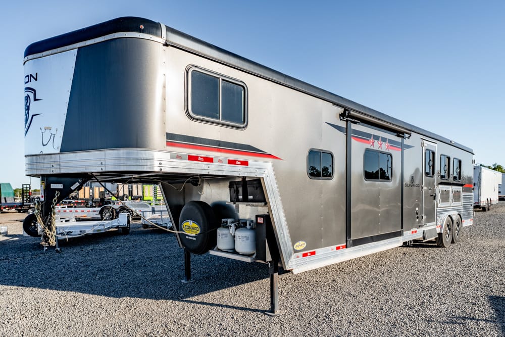 Large silver gooseneck horse trailer with red stripe, parked on gravel, propane tanks and spare tire visible at the front.