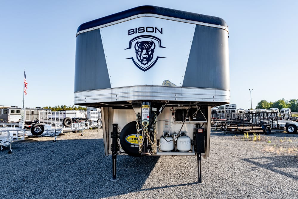 Front view of a Bison travel trailer elevated on its jack stands in a gravel trailer yard, with other trailers in the background and a blue sky.