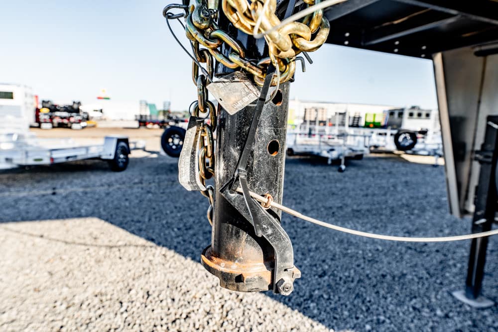 Close-up of a trailer hitch with safety chains and a padlock on a gravel lot; other trailers visible in the background.