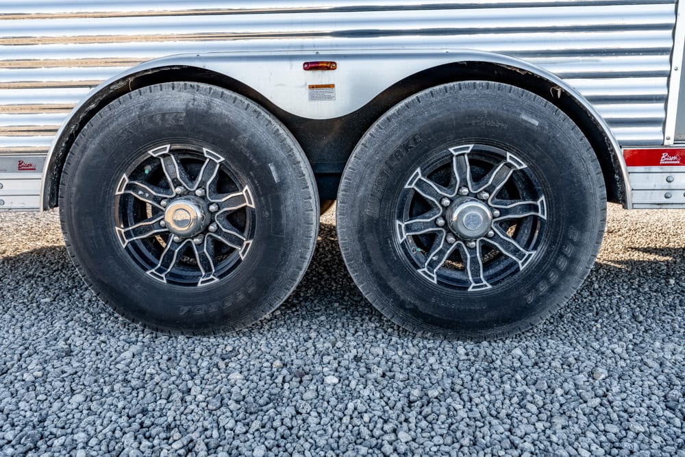 Dual-axle trailer with two large black tires and alloy wheels on a gravel surface under a silver trailer body.
