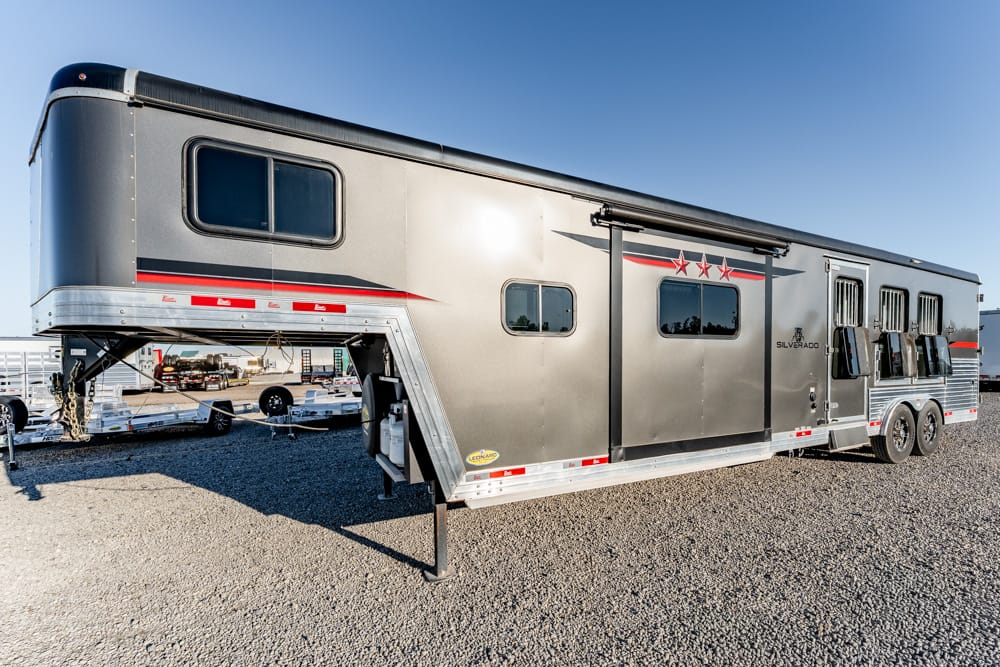 Long silver horse trailer parked on a gravel lot, with a three-star red graphic along the side and multiple windows