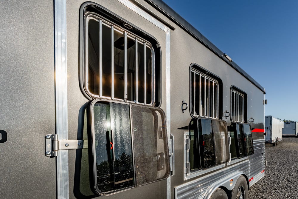 Side view of a gray horse trailer with barred windows and open hatch doors on a gravel lot, other trailers in the distance