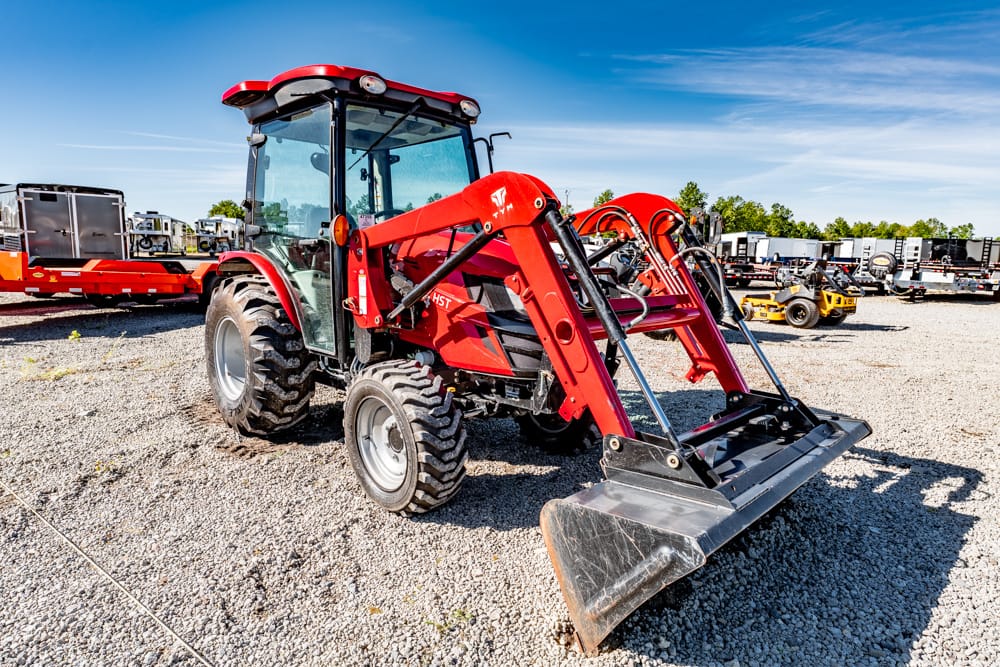 Red compact utility tractor with a hydraulic front-end loader and bucket on a gravel lot, blue sky above, other machinery in the background.