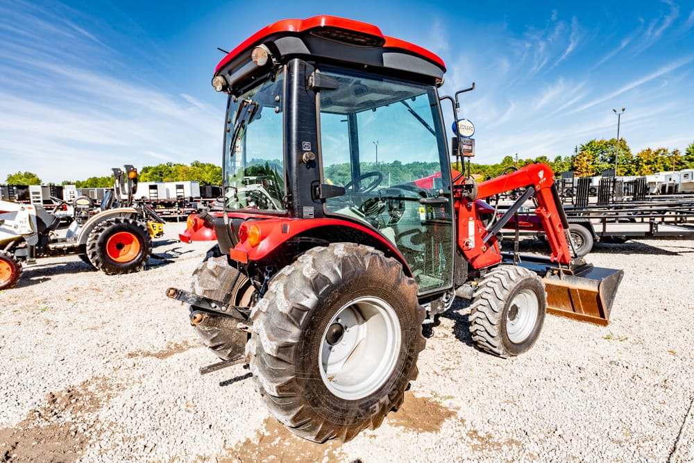 Red compact tractor with a front loader attachment parked on a gravel lot under a blue sky, with other equipment in the background