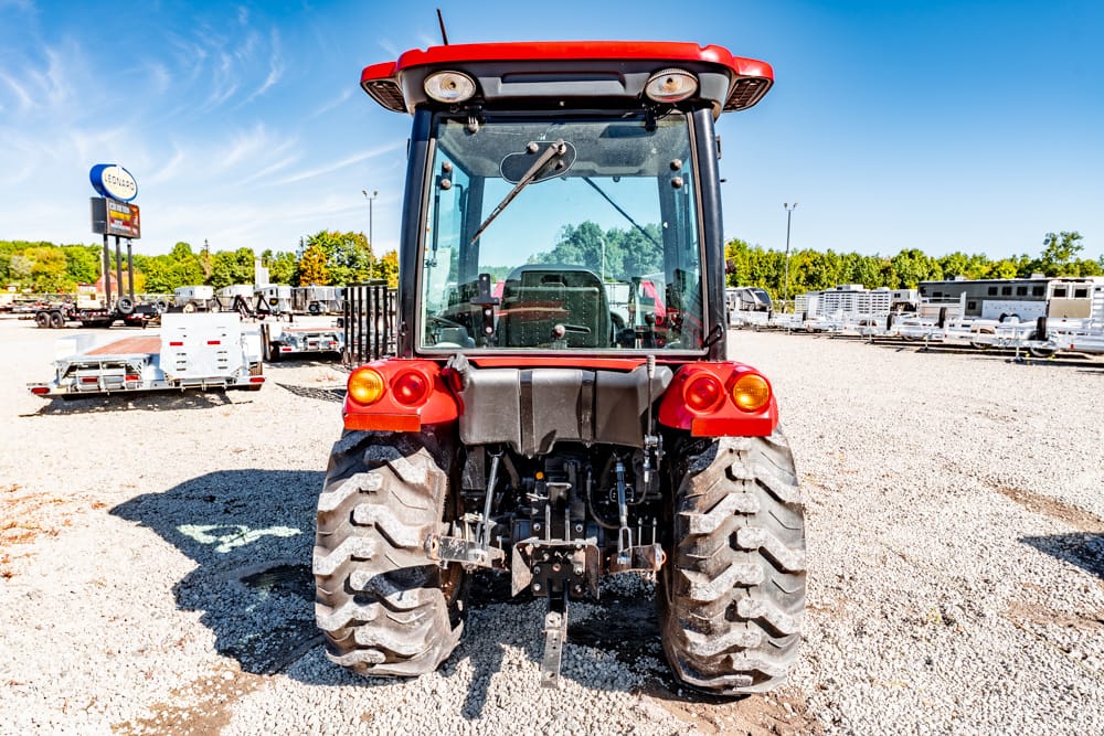 Rear view of a red tractor parked in a gravel lot with trailers and equipment in the background on a sunny day.