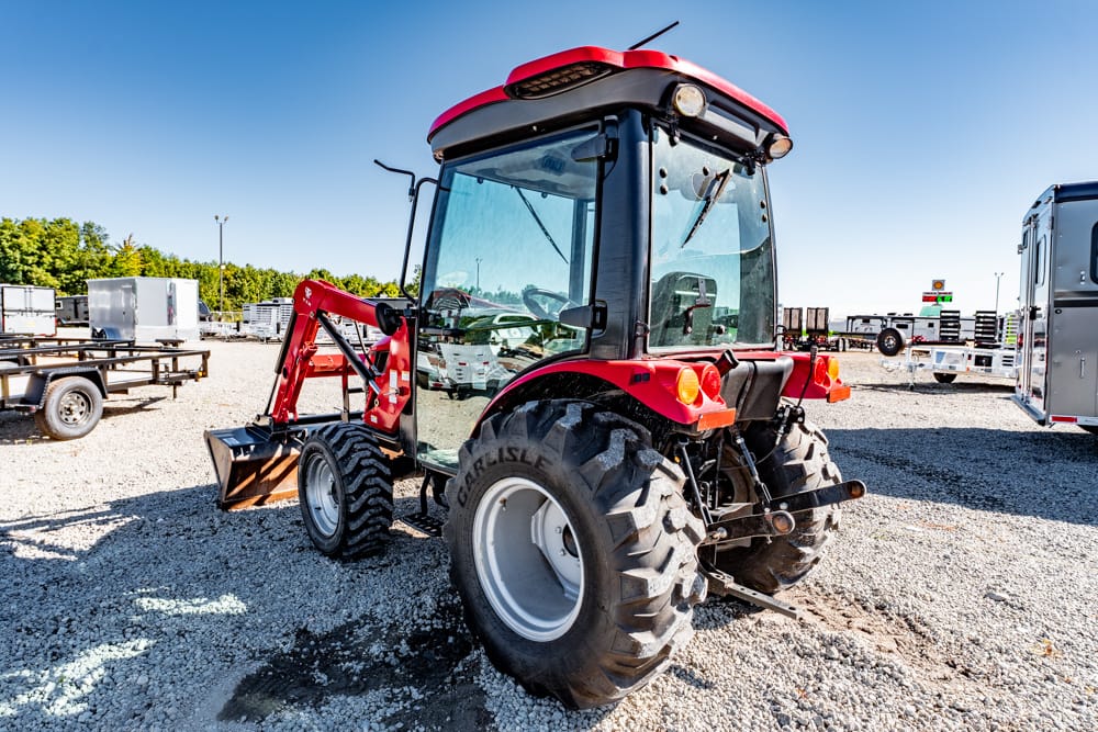 Red tractor with a front loader parked on gravel lot, surrounded by trailers and equipment under a clear blue sky.