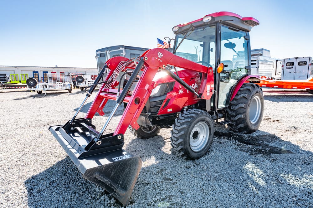 Red tractor with a front-end loader on a gravel lot at an equipment yard, with an American flag visible in the cab area.