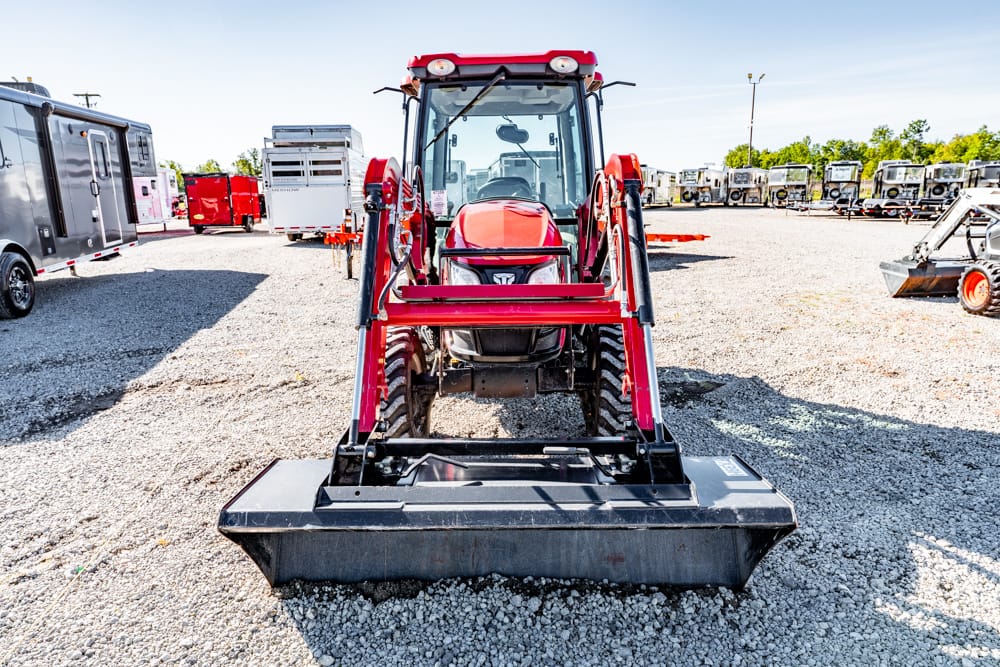 Front view of a red tractor with a large black front loader attachment in a gravel lot, with trailers and trucks in the background