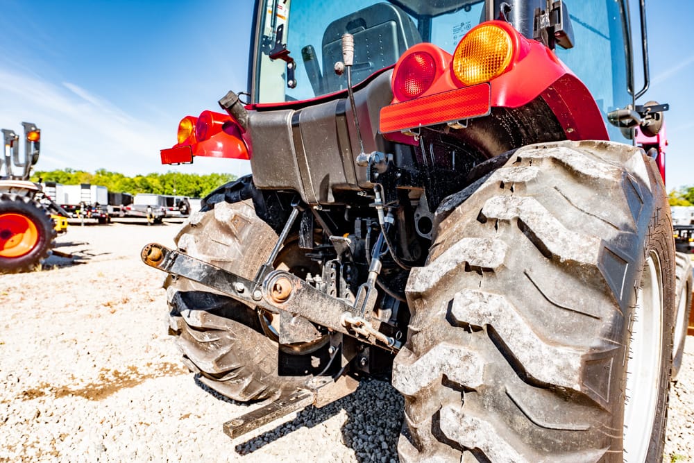 Rear view of a red tractor with oversized tires and a hitch on a gravel lot, with other equipment in the background.