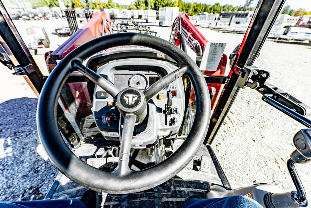 First-person view from inside a tractor cab, showing a large steering wheel, dashboard controls, and a gravel lot with other machinery outside.