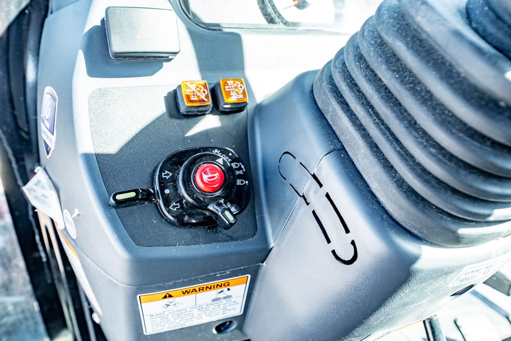 Control panel of heavy equipment showing a red emergency stop button, joystick, two orange toggle buttons, and a warning label.