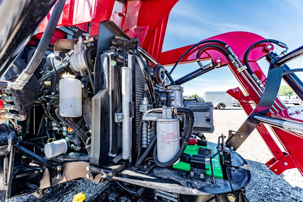 Close-up of a heavy machinery engine bay with hoses, filters, and radiator components exposed under a red arm/boom. This shows the mechanical setup of the equipment.