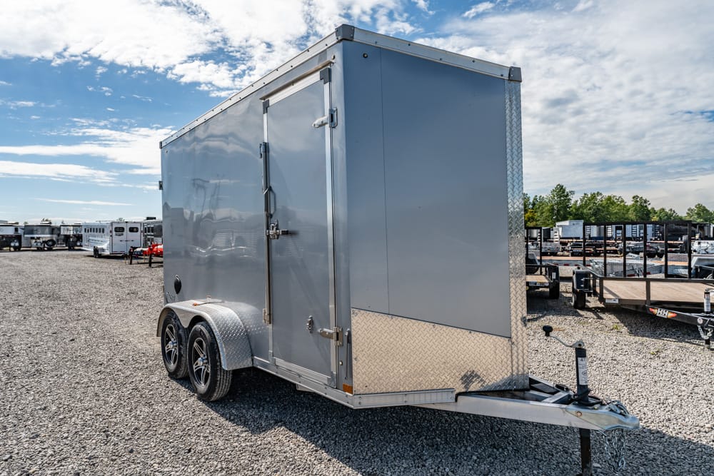 Silver enclosed cargo trailer with tandem wheels and diamond-plate front parked on a gravel lot among other trailers in a yard.