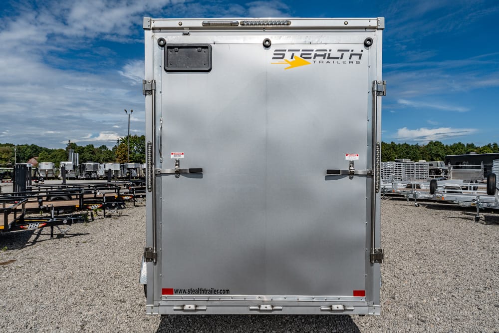 Back of a silver Stealth trailer with double doors and logo, parked in a gravel trailer lot on a sunny day.