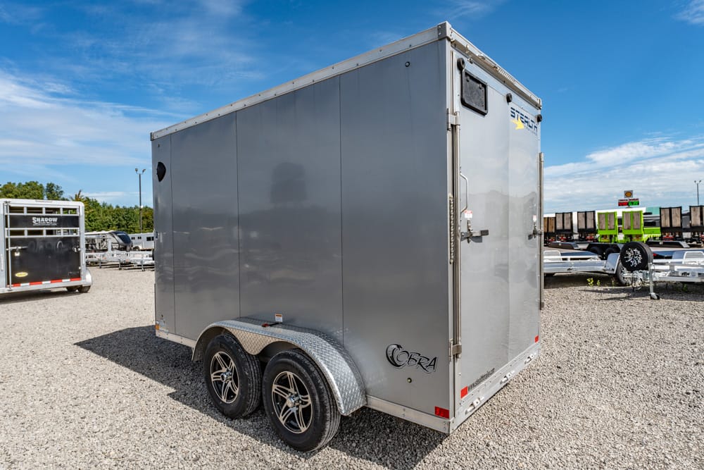 Gray enclosed cargo trailer parked on a gravel lot with dual axles and diamond-plate fenders, under a blue sky