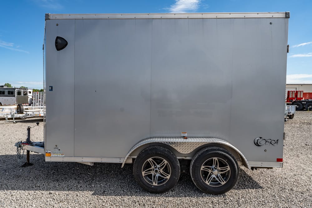 Gray enclosed cargo trailer with tandem wheels on a gravel lot; Cobra branding on the rear corner.