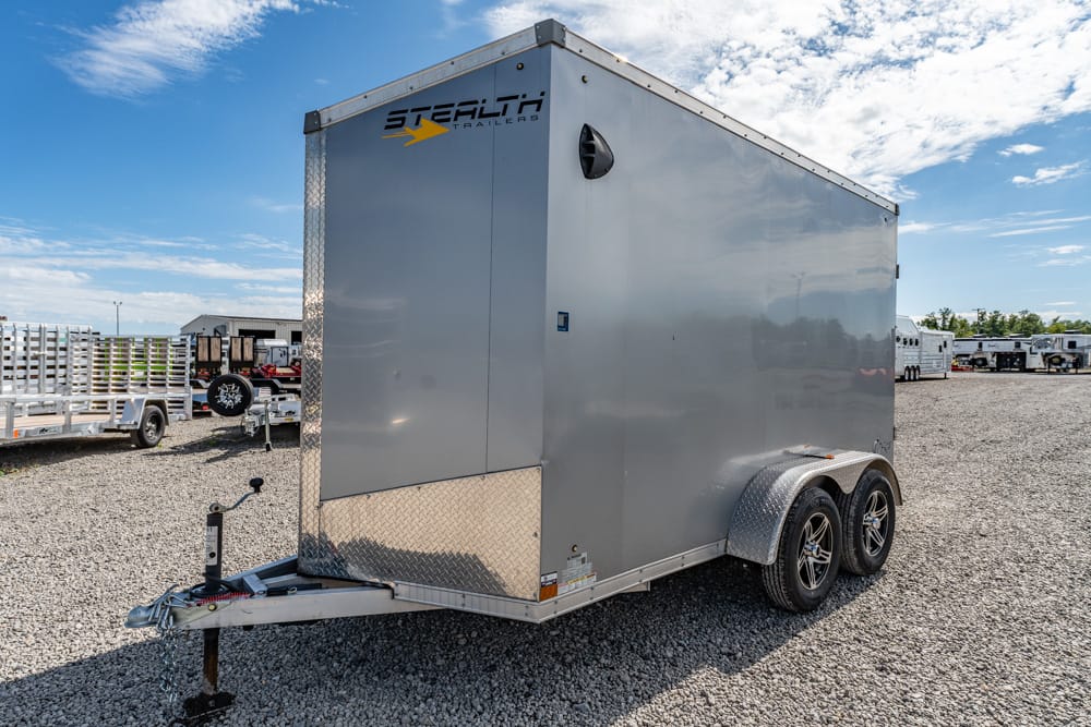 Gray enclosed cargo trailer with tandem axles and diamond-plate front on a gravel lot, under a blue sky with other trailers in the background, logo reads Stehlit Trailers.
