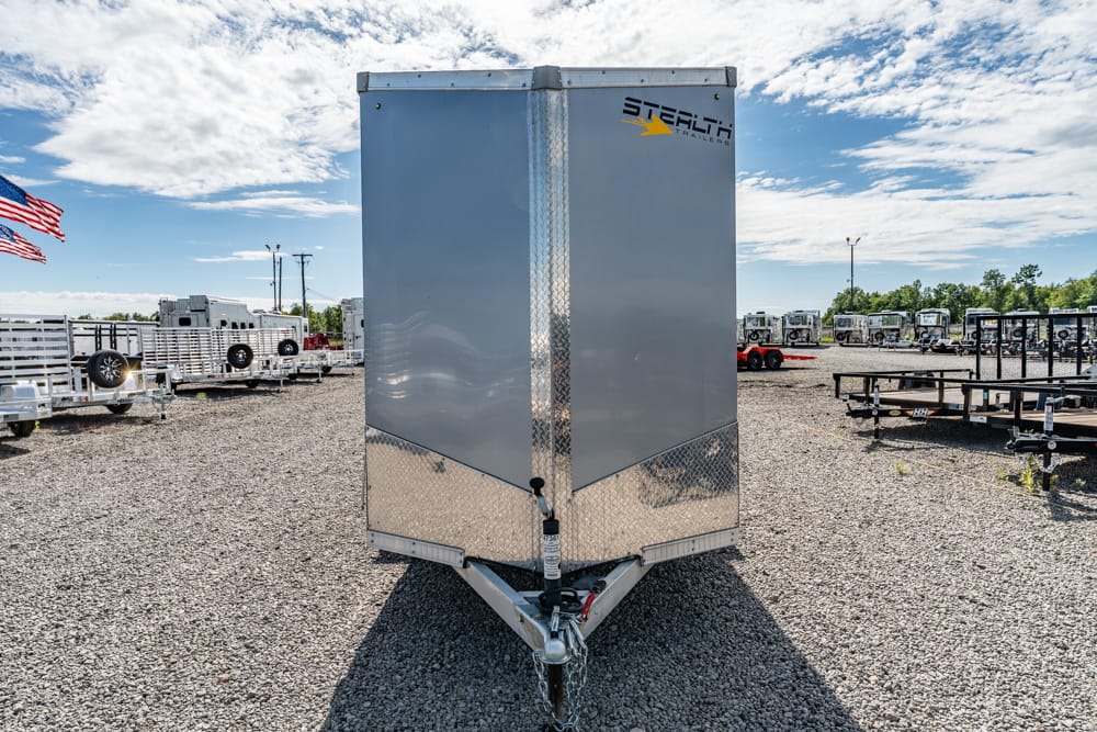 Front view of a tall silver enclosed trailer on a gravel lot, with other trailers in the background and a logo in the upper right corner.