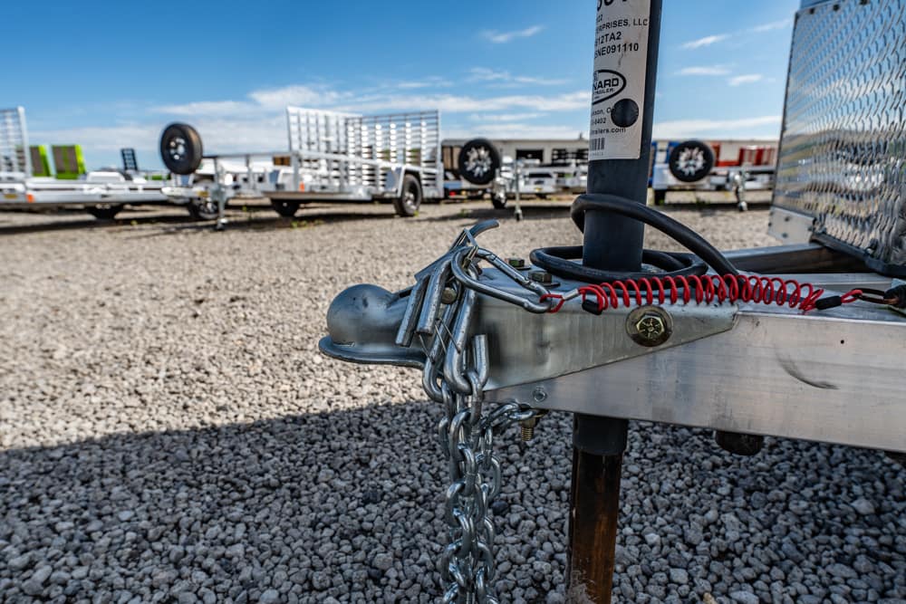 Close-up of a trailer hitch with safety chains and a red coiled cable on a metal frame, with a gravel lot and trailers in the background.