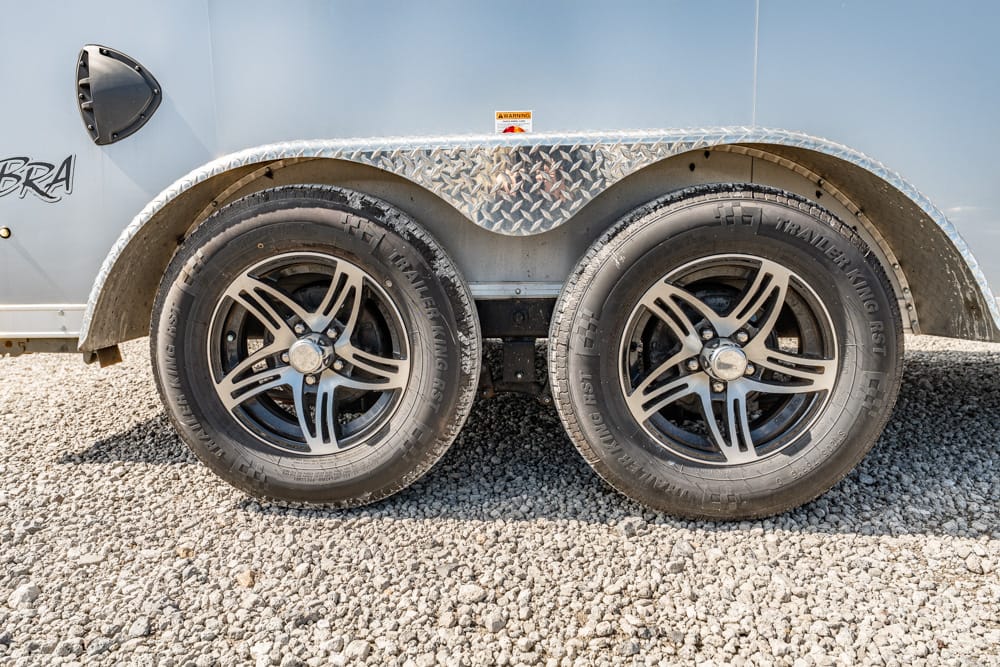Dual trailer wheels with chrome rims on a gravel lot under a diamond-plate fender on a blue trailer surface.