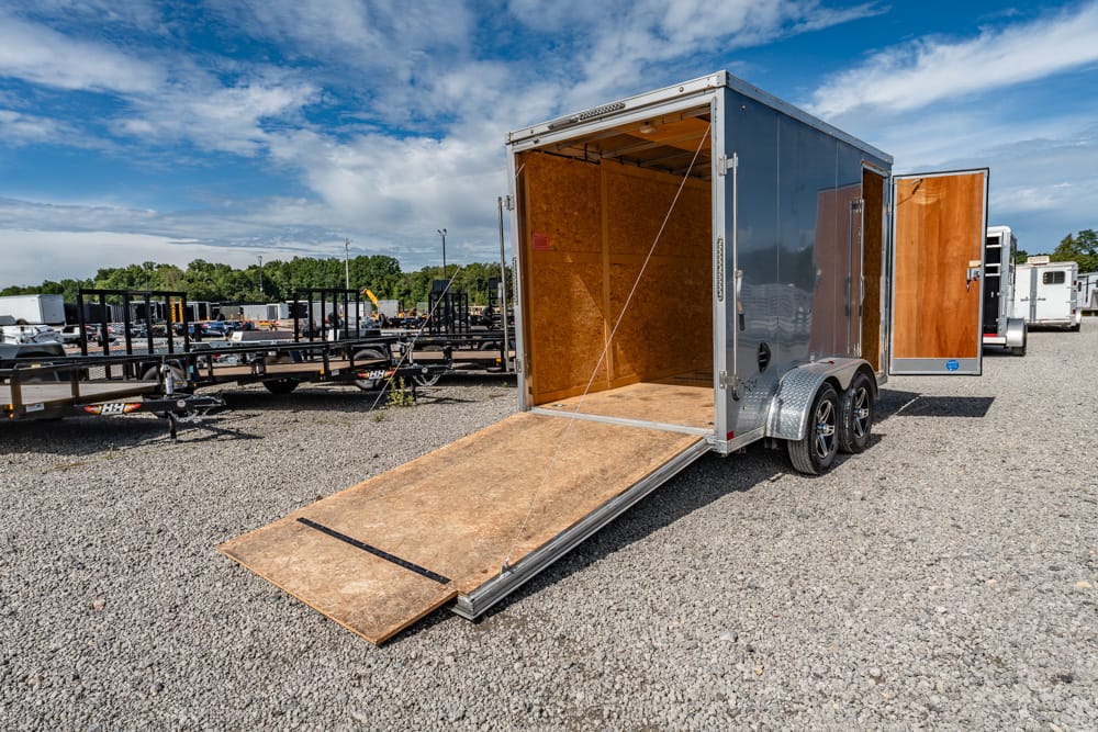 Silver enclosed cargo trailer with the rear ramp down, exposing a plywood interior on a gravel lot with other trailers nearby under a blue sky.