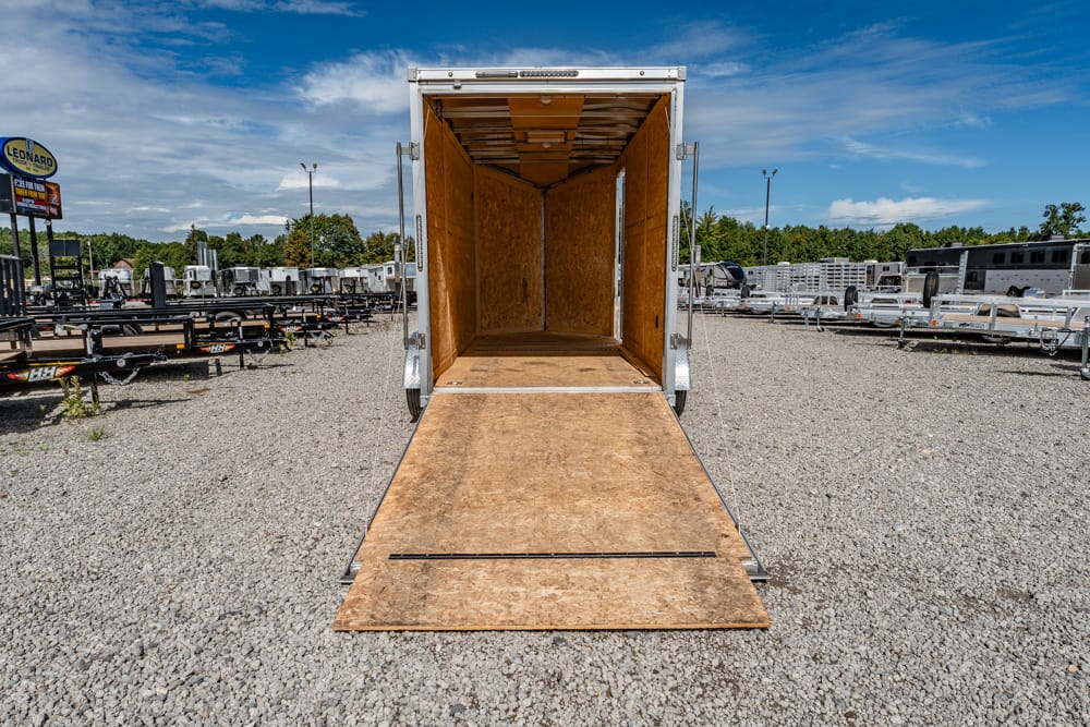 Open cargo trailer with wooden interior and lowered ramp on a gravel lot, surrounded by other trailers and equipment in a parking area.
