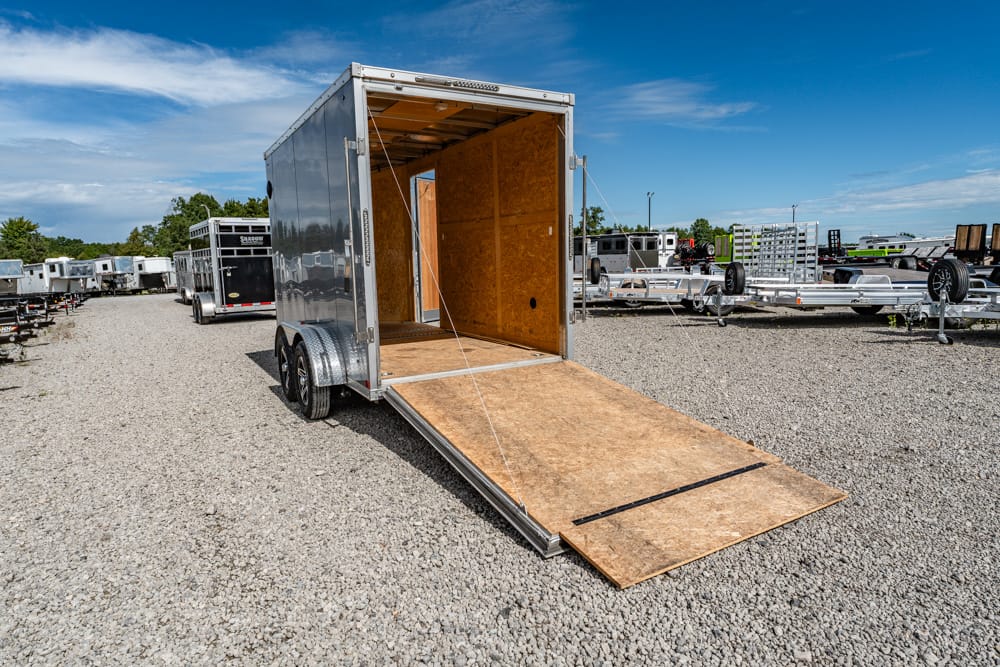 Silver cargo trailer with its rear ramp lowered onto a gravel lot, empty interior visible, other trailers in the background.