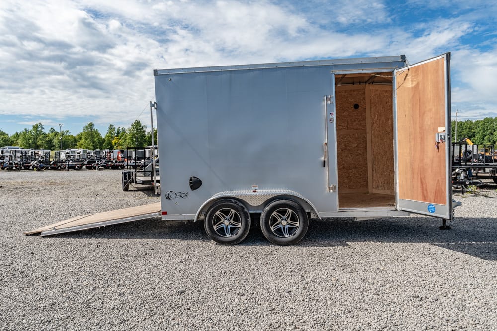 Silver enclosed cargo trailer with open rear door and lowered ramp on a gravel lot, ready for loading.