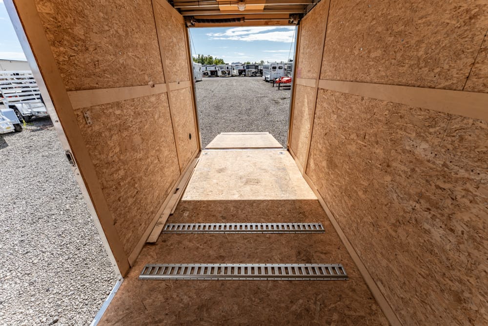 Interior of an empty plywood-walled cargo trailer with a raised ramp leading to a gravel lot and other trailers in the distance