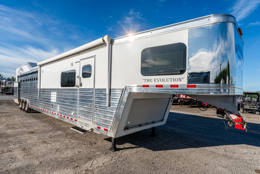 White fifth-wheel trailer with chrome aluminum siding parked on a gravel lot under a blue sky, labeled 'The Evolution' on the front.