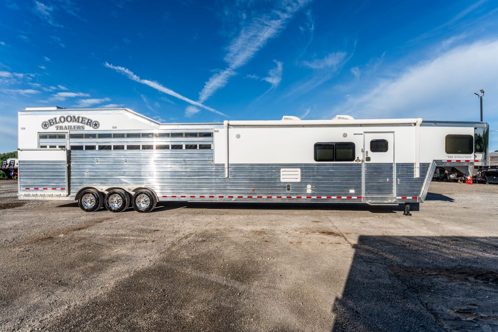 Long silver aluminum horse trailer with three axles and red-and-white reflective tape parked on a dirt lot under a bright blue sky.