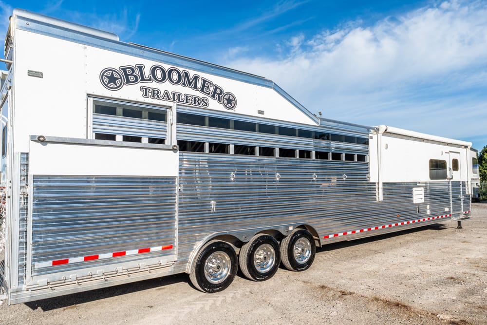 Silver Bloomer Trailers horse trailer with triple axles parked on gravel under a blue sky, logo visible on upper front panel.