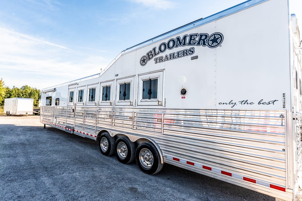 White Bloomer horse trailer with chrome wheels and multiple windows, parked on a gravel lot.