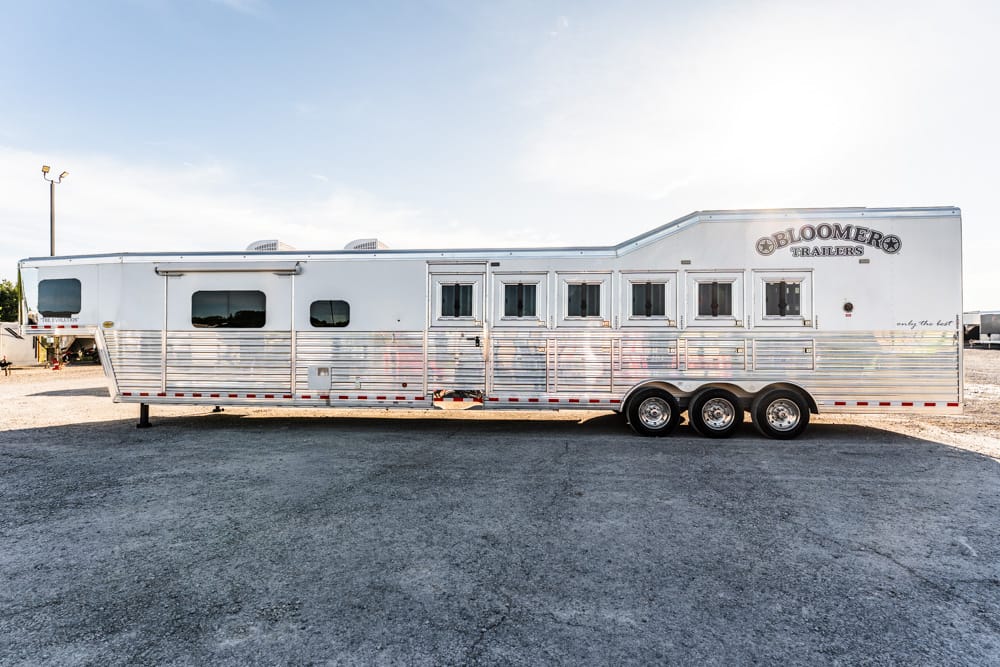 Long white Bloomer horse trailer with multiple windows parked on gravel outside, triple-axle design visible.