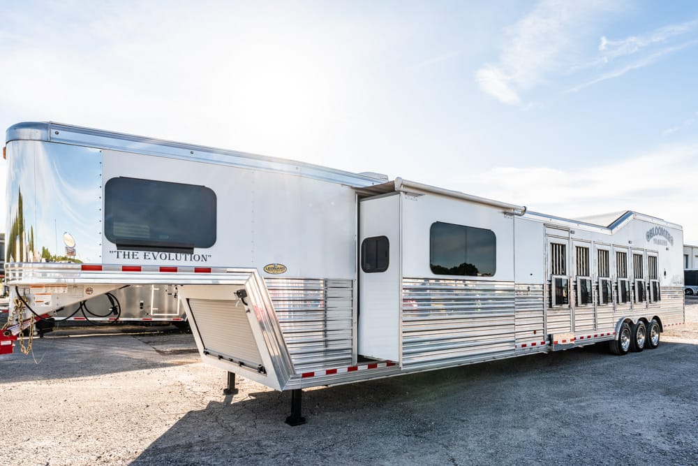 White multi-section horse trailer with chrome panels and an open ramp at a gravel lot.