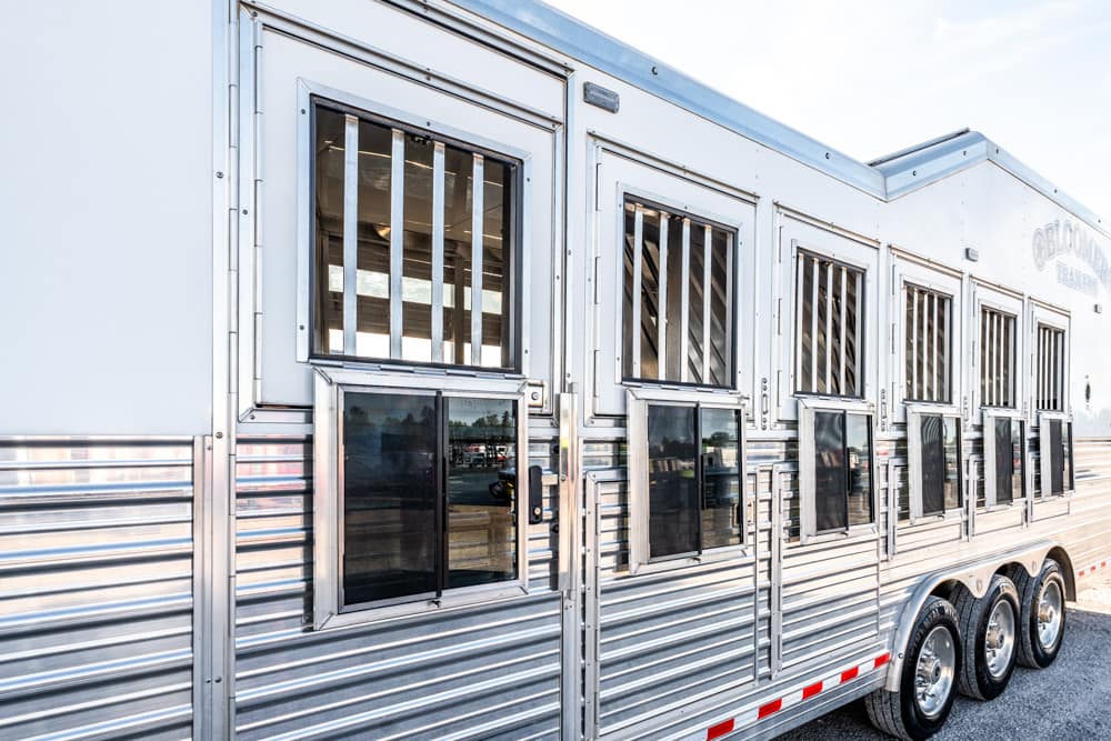 Shiny aluminum livestock trailer with barred vents and multiple windows parked on a gravel lot.