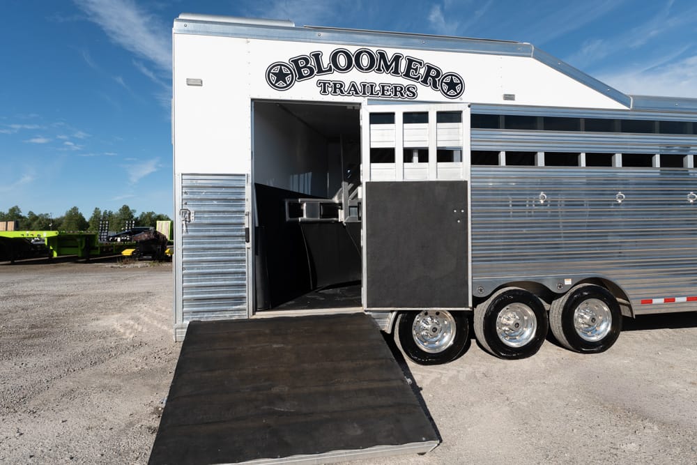 Silver Bloomer Trailers horse trailer with ramp down, parked on a gravel lot under a blue sky.