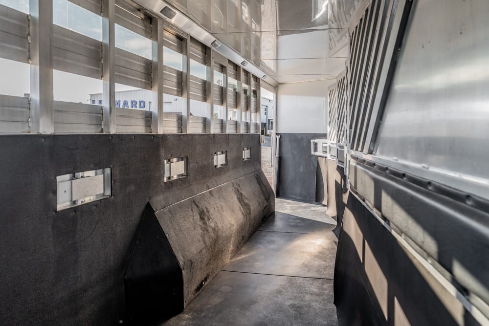 Interior of a metal livestock trailer with a narrow aisle, windows along the left wall, and dark padded sides leading to a bright doorway at the end