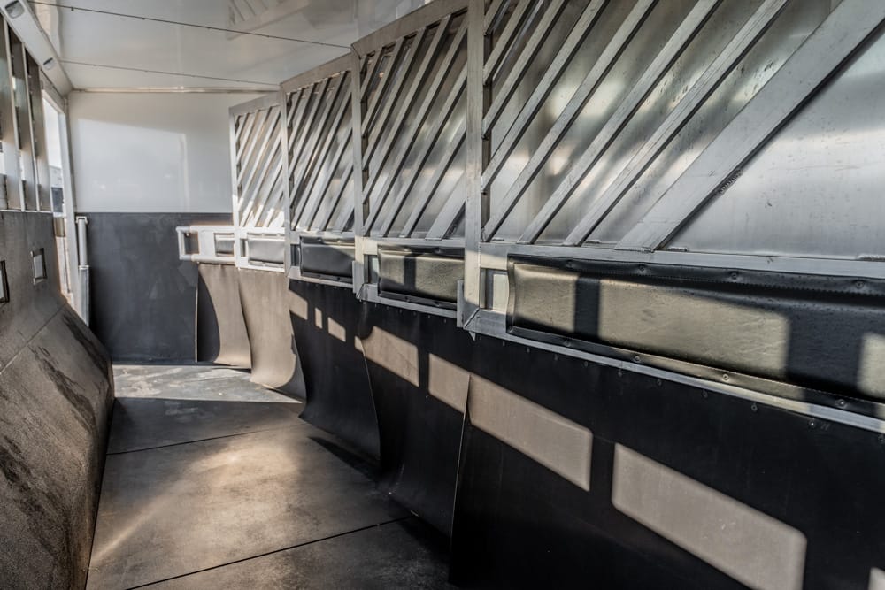Interior of a metal livestock trailer with diagonal wall grates, empty stalls, and dark rubberized lower panels with sunlight patterns on the floor.