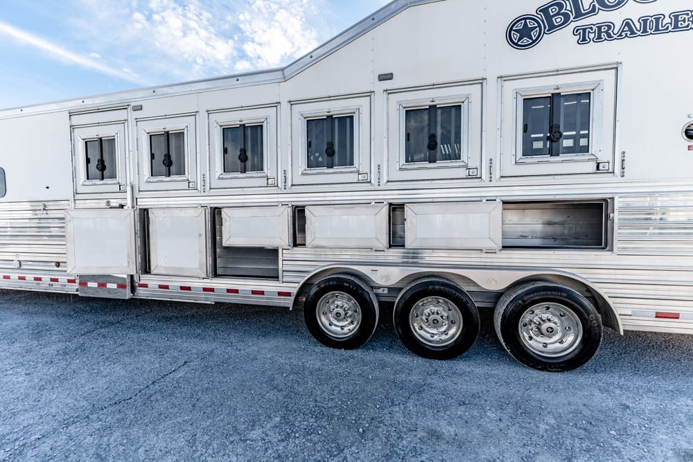 Silver aluminum horse trailer with multiple square windows and side storage compartments, parked on gravel.
