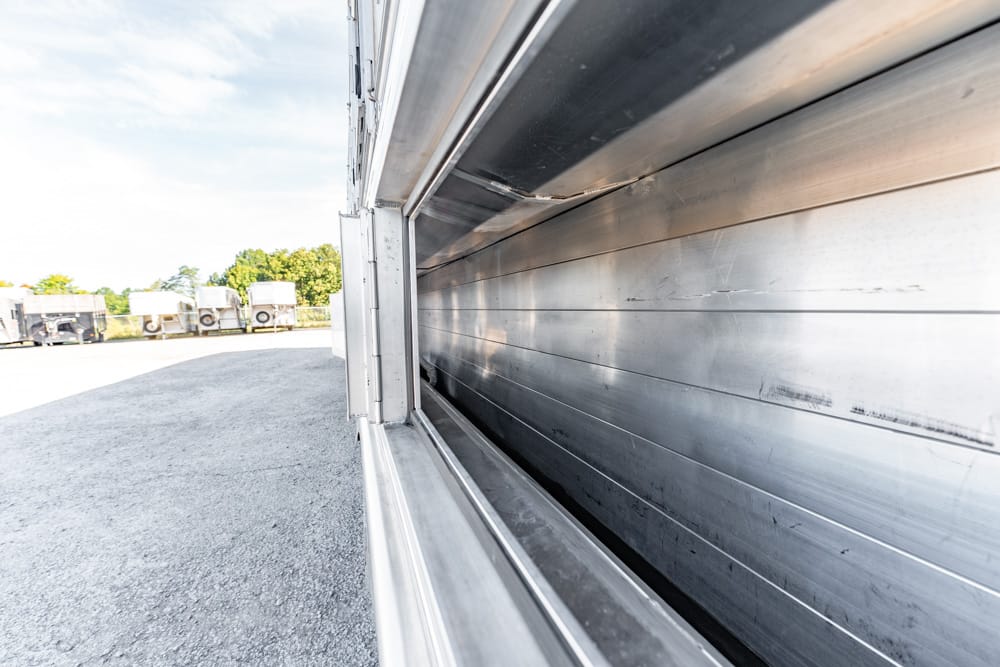 Close-up of a metal truck trailer with a rolled-down side door, docked in an outdoor lot with other trailers in the distance