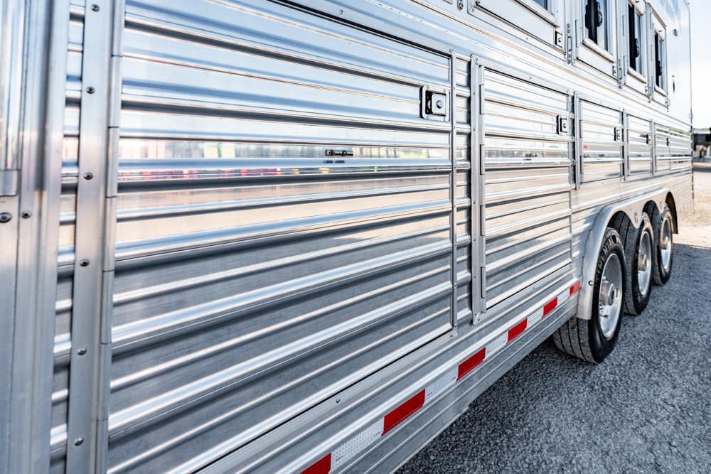 Side view of a shiny aluminum trailer with ribbed panels and multiple wheels on a gravel surface