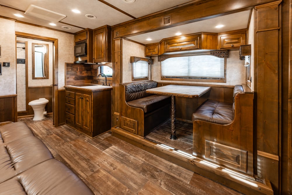 Wood-paneled RV interior with a dinette booth, small kitchen (sink and stove), upper cabinets, and a peek at the bathroom in the background.