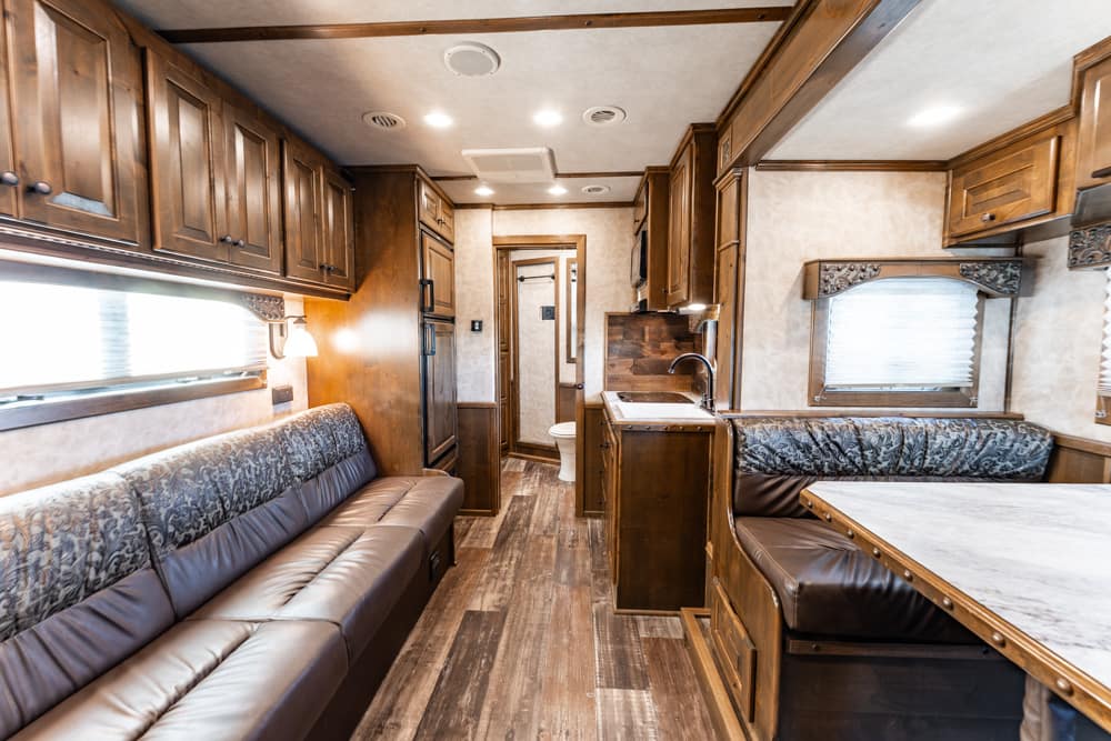 Interior of an RV living area with dark wood cabinets, a long leather sofa, and a booth-style dining table with a marble-like top on the right.