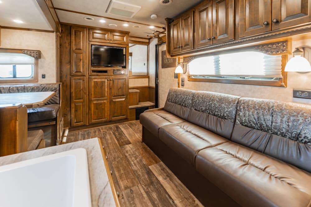 Interior of a luxurious RV living area with a brown leather sofa, dark wood cabinets, and a mounted flat-screen TV across from the seating area