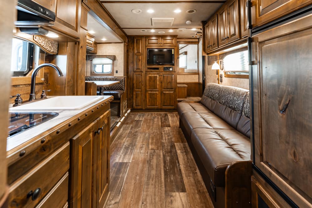 Interior of a wooden RV kitchen and living area with sink, stove, and a long brown leather couch facing a TV cabinet at the back.