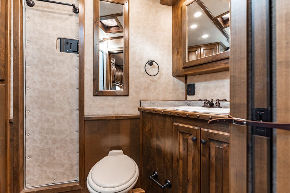RV bathroom with wooden vanity, marble countertop, mirror, and white toilet on beige walls with warm lighting and storage cabinets.