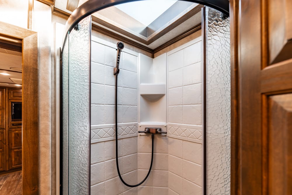 Shower stall in a wood-paneled bathroom with white tiled walls, a handheld showerhead, and a curved glass door framed by frosted glass panels.