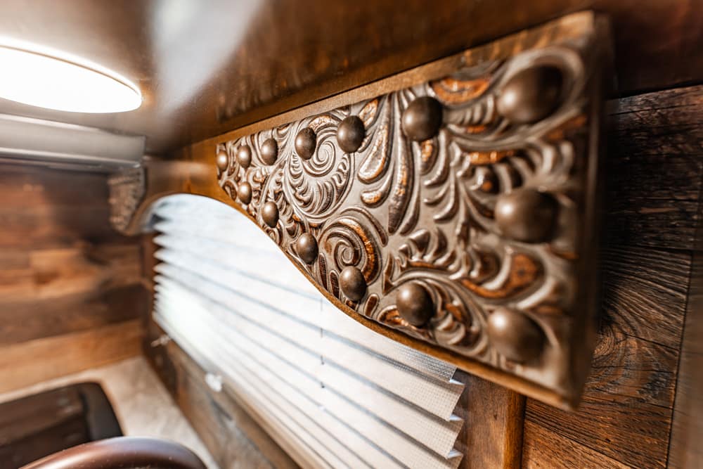 Ornate carved wooden panel with scrolling floral pattern and round metal studs against a rustic wooden interior near a window with blinds.